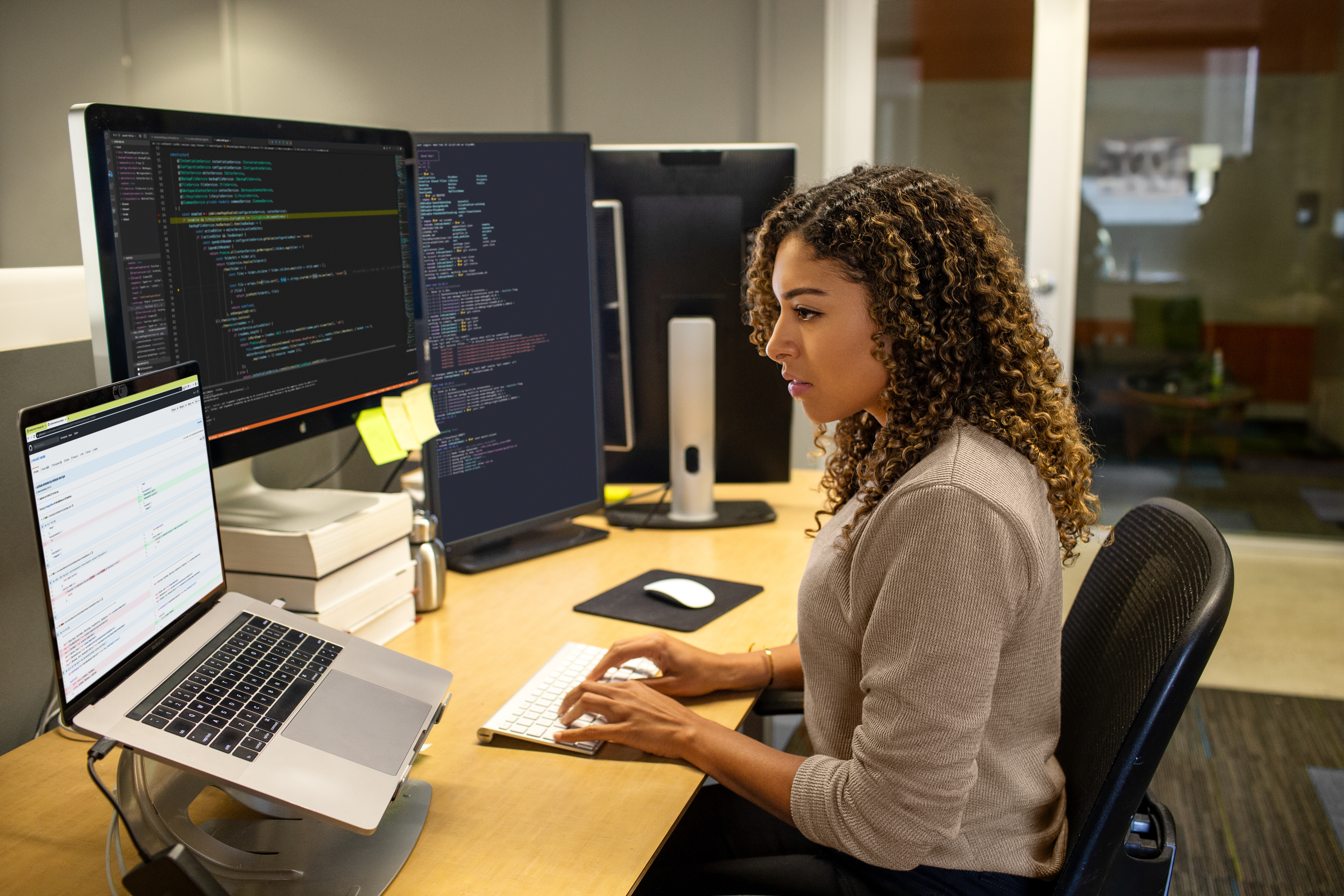Real people, real offices. Black female developer working at enterprise office workspace. Focused work. She has customized her workspace with a multi-monitor set up. Women who code, women developers, women engineers, code, develop, Black developer, engineer, Visual Studio, Azure.